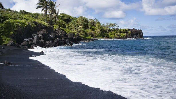 A beach with dark sand, ocean waves, and lush greenery with palm trees. - Family Getaway 3BD Across from  Beach-Pacific Shores B102 (Kihei)