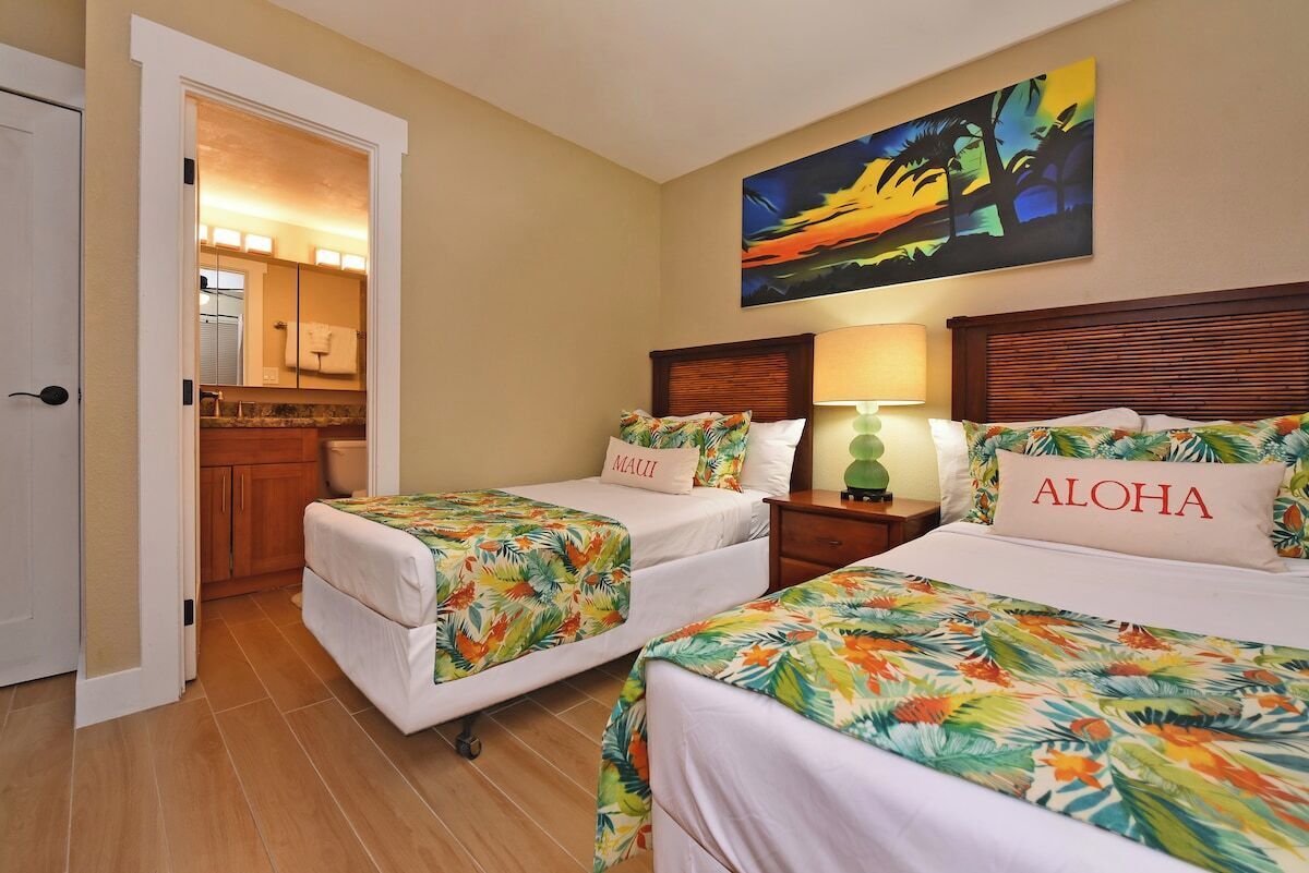 A hotel room with two beds, each with a floral bedspread and pillows labeled "Maui" and "Aloha". There is a painting of a sunset on the wall, and a bathroom is visible through an open door.