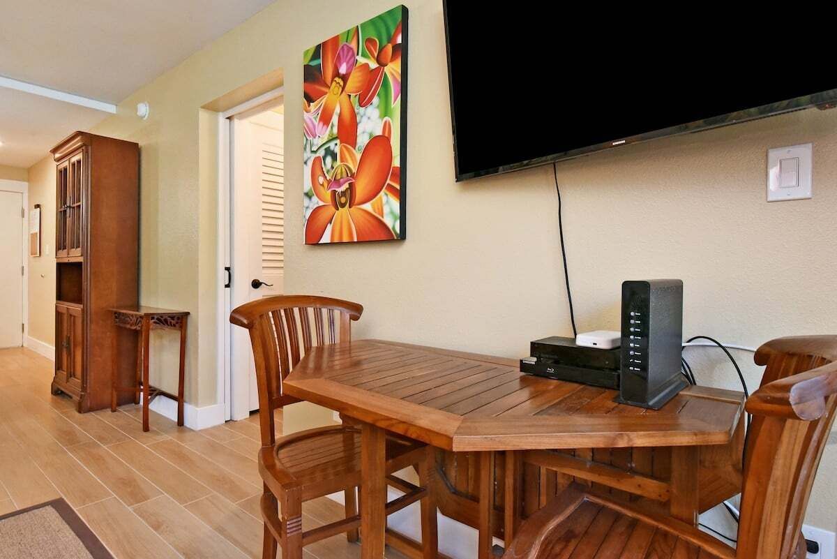 A dining area with a wooden table and chairs, a painting on the wall, and a television mounted above the table.