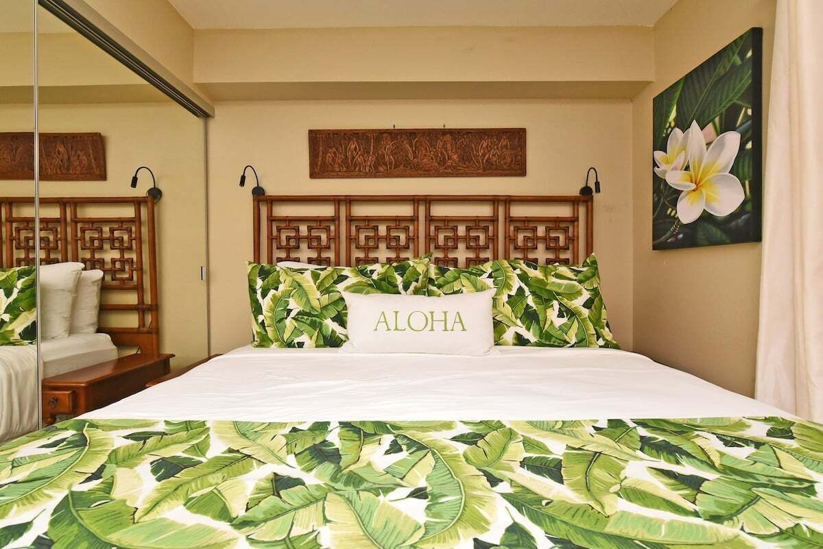 A hotel room with a bed featuring a tropical leaf pattern bedspread, a wooden headboard with a geometric design, a pillow with the word "ALOHA," and a wall-mounted artwork of a flower.