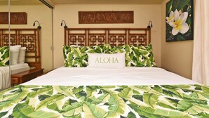 A hotel room with a bed featuring a tropical leaf pattern bedspread, a wooden headboard with a geometric design, a pillow with the word "ALOHA," and a wall-mounted artwork of a flower. - Family Getaway 3BD Across from  Beach-Pacific Shores B102 (Kihei)