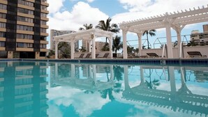 Une piscine dotée de chaises longues et d’une pergola, entourée d’immeubles imposants et de palmiers.
