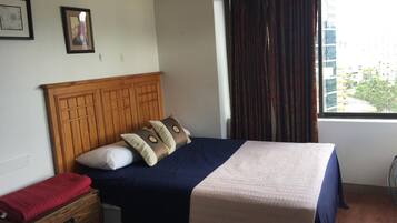 Una cama bien hecha con tablero de madera, dos almohadas y una manta azul y beige. Se aprecia una mesita de noche con una toalla roja, un cuadro enmarcado en la pared y una ventana con cortinas.