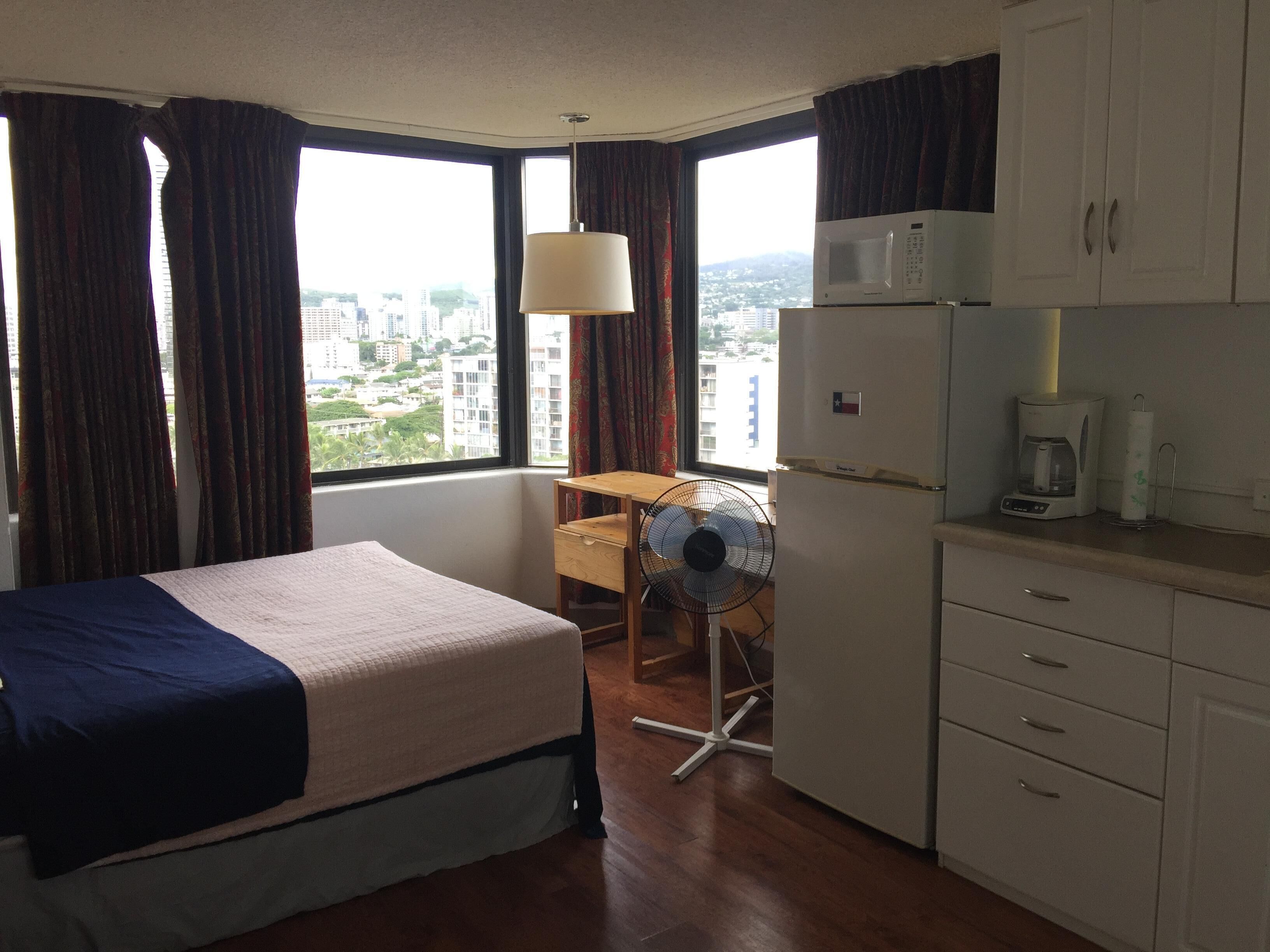 Habitación de hotel con cama, escritorio, ventilador, microondas, cafetera y vistas a la ciudad.