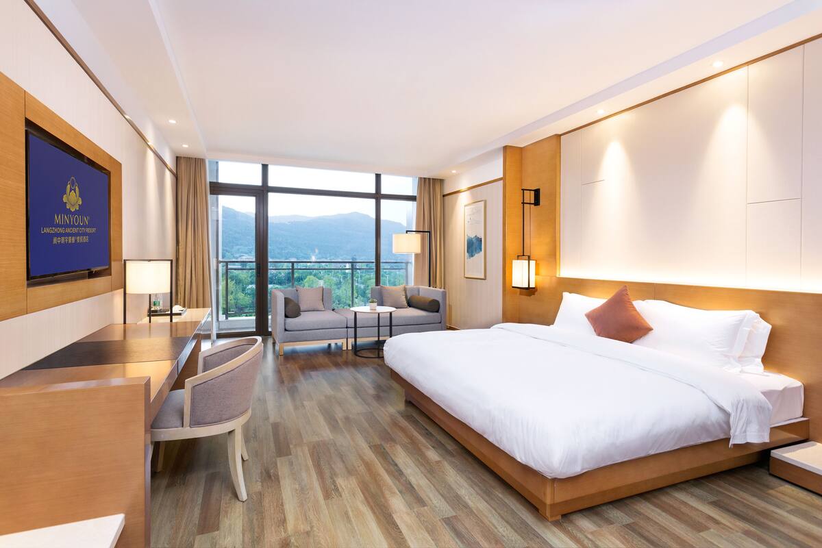 A hotel room with a large bed, a desk, a sofa, and a TV. There is a view of mountains through the window.