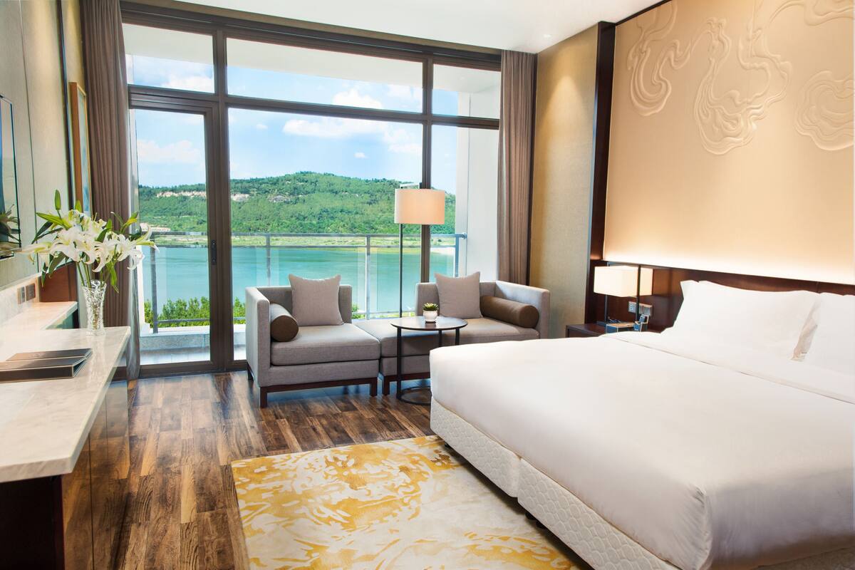 A hotel room with a large bed, a sofa, a small table, and a view of a lake and hills.