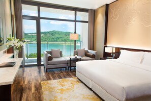 Quarto de hotel com uma cama grande, um sofá, uma mesinha e vista para um lago e colinas.