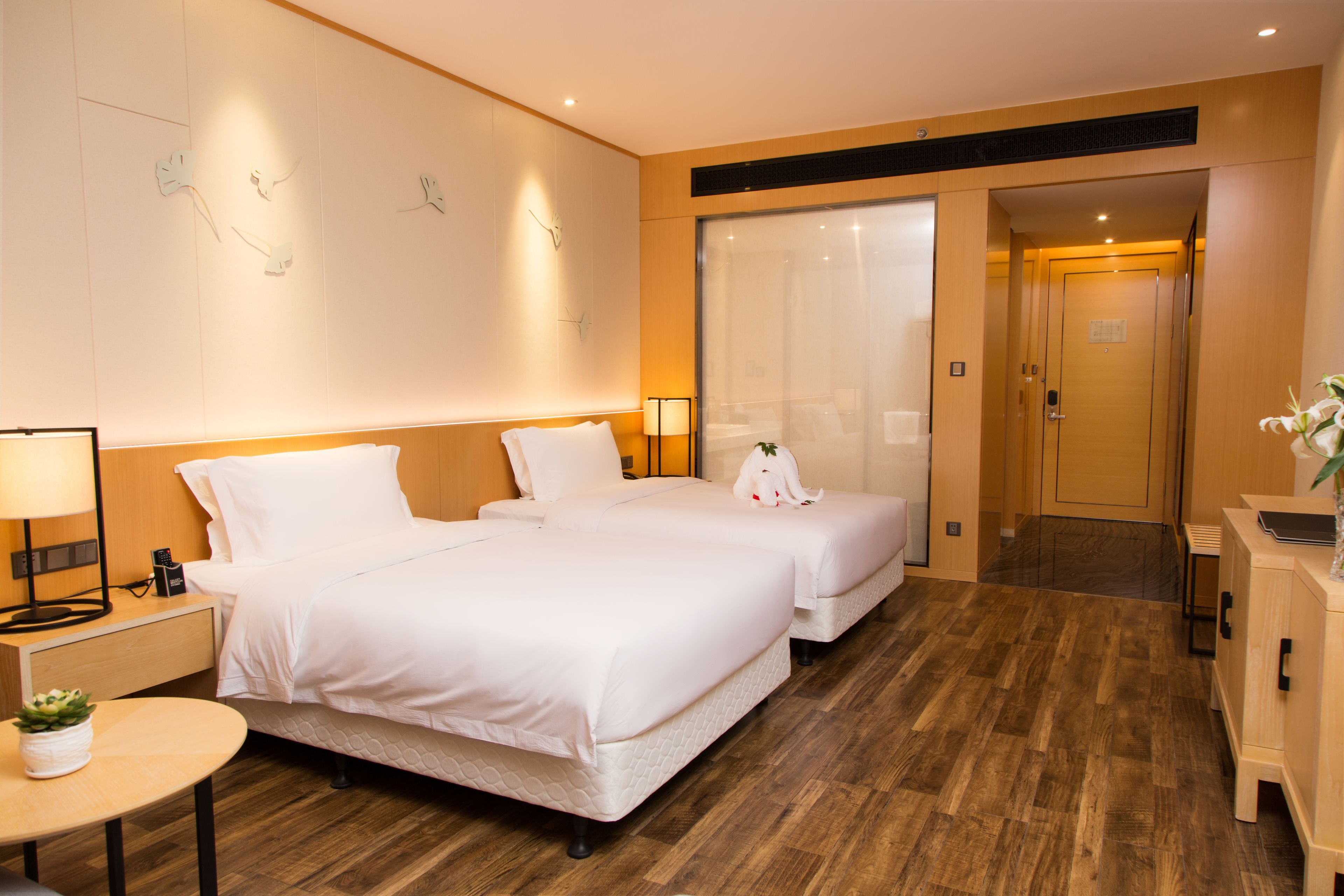 A hotel room with two beds, a bedside table with a lamp, a small table with a plant, and a wooden floor.