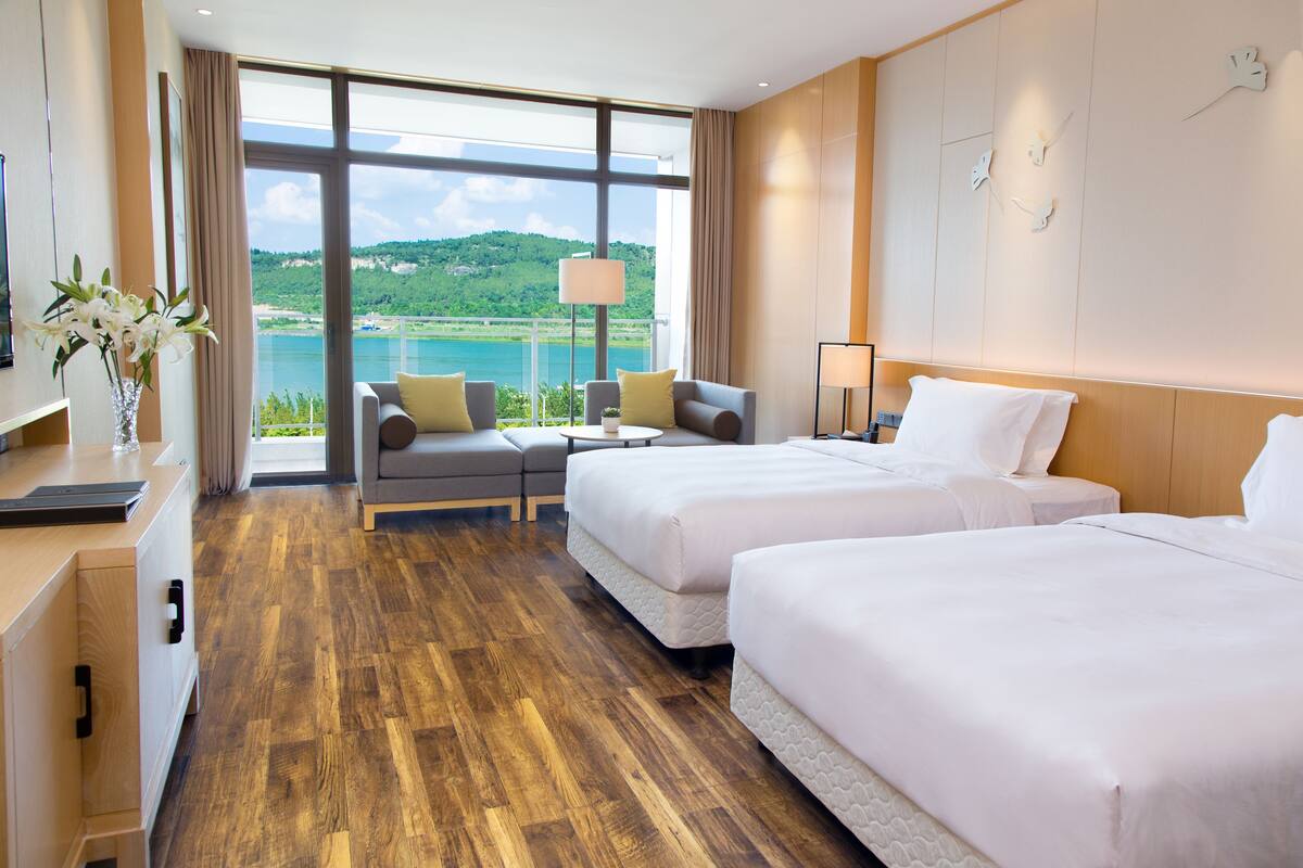 A hotel room with two beds, a sofa, a desk, and a large window offering a view of a lake and hills.