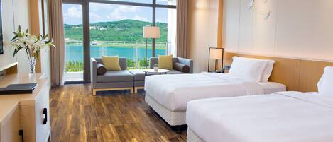 A hotel room with two beds, a sofa, a desk, and a large window offering a view of a lake and hills.