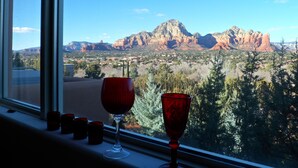 Una ventana con una copa de vino tinto y dos vasos rojos en el alféizar, con vistas a un paisaje montañoso y cielo despejado.