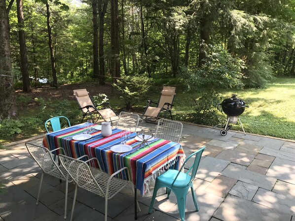 An outdoor patio with a table set for dining, surrounded by chairs, and a barbecue grill in the background.