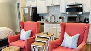 A compact living space with two red armchairs, a small coffee table, and two woven ottomans. A kitchenette with white cabinets and stainless steel appliances is visible in the background. - Studio Near the Plantations (Charleston)