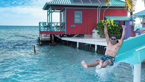 Eine Person gleitet von einer Rutsche, die an ein rotes Gebäude mit einer Veranda angebaut ist, in türkisblaues Wasser.