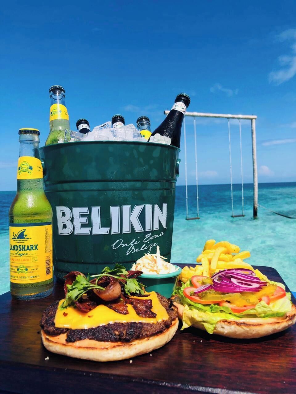 A beachside dining setup with a Belikin cooler, two burgers, a salad, and a drink.