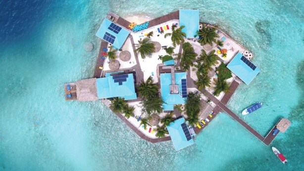 Eine Vogelperspektive zeigt einen tropischen Inselresort mit mehreren Schwimmbecken, Palmen und einer Anlegestelle mit Booten.