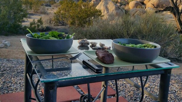 Une table en verre sur laquelle sont disposés un steak et deux salades, le tout installé à l'extérieur, avec un paysage rocheux et une végétation clairsemée en arrière-plan.