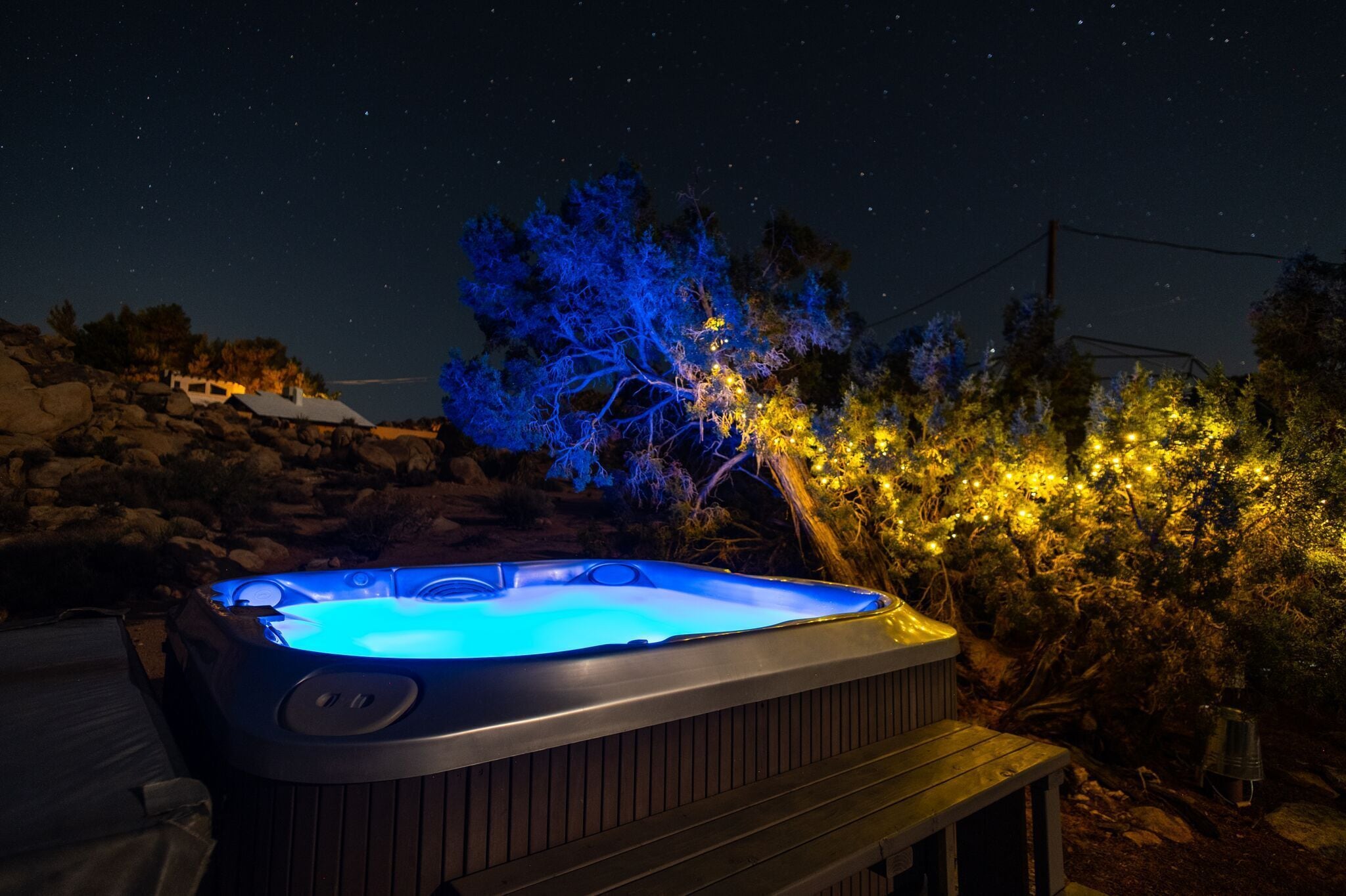 A hot tub with a wooden deck, surrounded by rocks and trees, under a night sky with stars.
