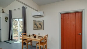 Un coin repas avec une table dressée pour deux personnes, une fenêtre avec des rideaux, un appareil de climatisation et une porte rouge.