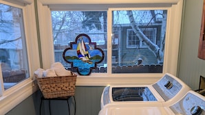 A laundry room with a large washer and dryer, a window with a sailboat stained glass, a wicker basket with towels, and a colorful rug on the floor. - Walking distance everywhere, yard, front porch and hot tub! (Coeur d'Alene)