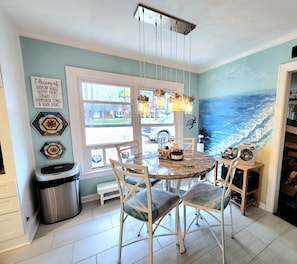 A dining area with a round table, chairs, a trash can, and a window with a view of the outside.