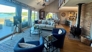 A spacious living room with a large window overlooking a scenic view, a cozy seating area, and a kitchen area visible through an open doorway. - Oceanfront - 3 Acre Private Beach Property, See Whales & Eagles from the Deck (Eastport)