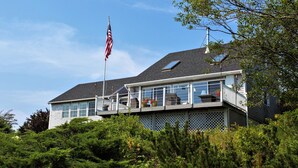 A two-story house with a balcony, a flagpole with an American flag, and a view of the surrounding greenery. - Oceanfront - 3 Acre Private Beach Property, See Whales & Eagles from the Deck (Eastport)