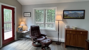 A room with a red door, a window with a view of trees, a leather chair, a lamp, a framed picture, and a cabinet with a bowl. - The Real Vermont All Seasons Get-Away Home - Part of Queche Lakes Country Club (Quechee)