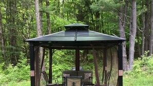 Ein Pavillon mit Tisch und Stühlen, umgeben von einer rot gemusterten Decke, eingebettet in einem Waldgebiet.