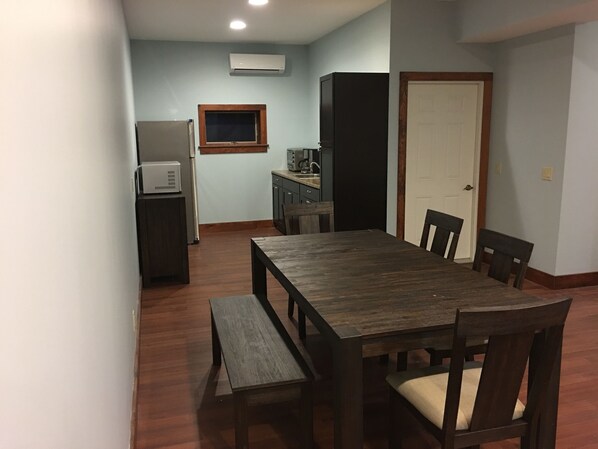 A dining area with a wooden table and chairs, a small kitchen area with a refrigerator and microwave, and a door leading to another room.