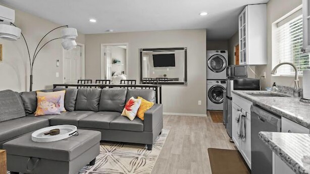 Un appartement moderne avec un canapé gris, un coin repas, une cuisine équipée d’un lave-linge et d’un sèche-linge intégrés, et une grande fenêtre.