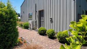 Il s’agit d’un bâtiment moderne à l’extérieur gris, doté d’une porte et d’une petite fenêtre. Un chemin pavé mène à l’entrée, et la zone est entourée de verdure.