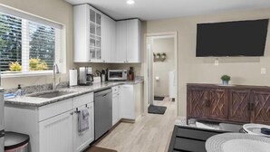 Une cuisine compacte avec des meubles blancs, un four à micro-ondes et un évier. Il y a un petit coin repas avec une table et des chaises. Un téléviseur est fixé au mur, et il y a une fenêtre avec des stores.