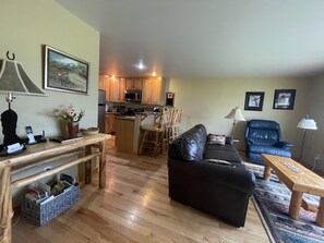 A living room with a black leather sofa, wooden coffee table, and a dining area with a wooden table and chairs. A kitchen with wooden cabinets and a microwave is visible in the background.