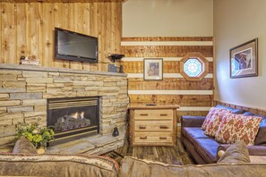 Un salon chaleureux avec une cheminée en pierre, un téléviseur à écran plat fixé au-dessus, un mur en bois orné d’un vitrail hexagonal, et un canapé en cuir marron agrémenté de coussins à motifs.