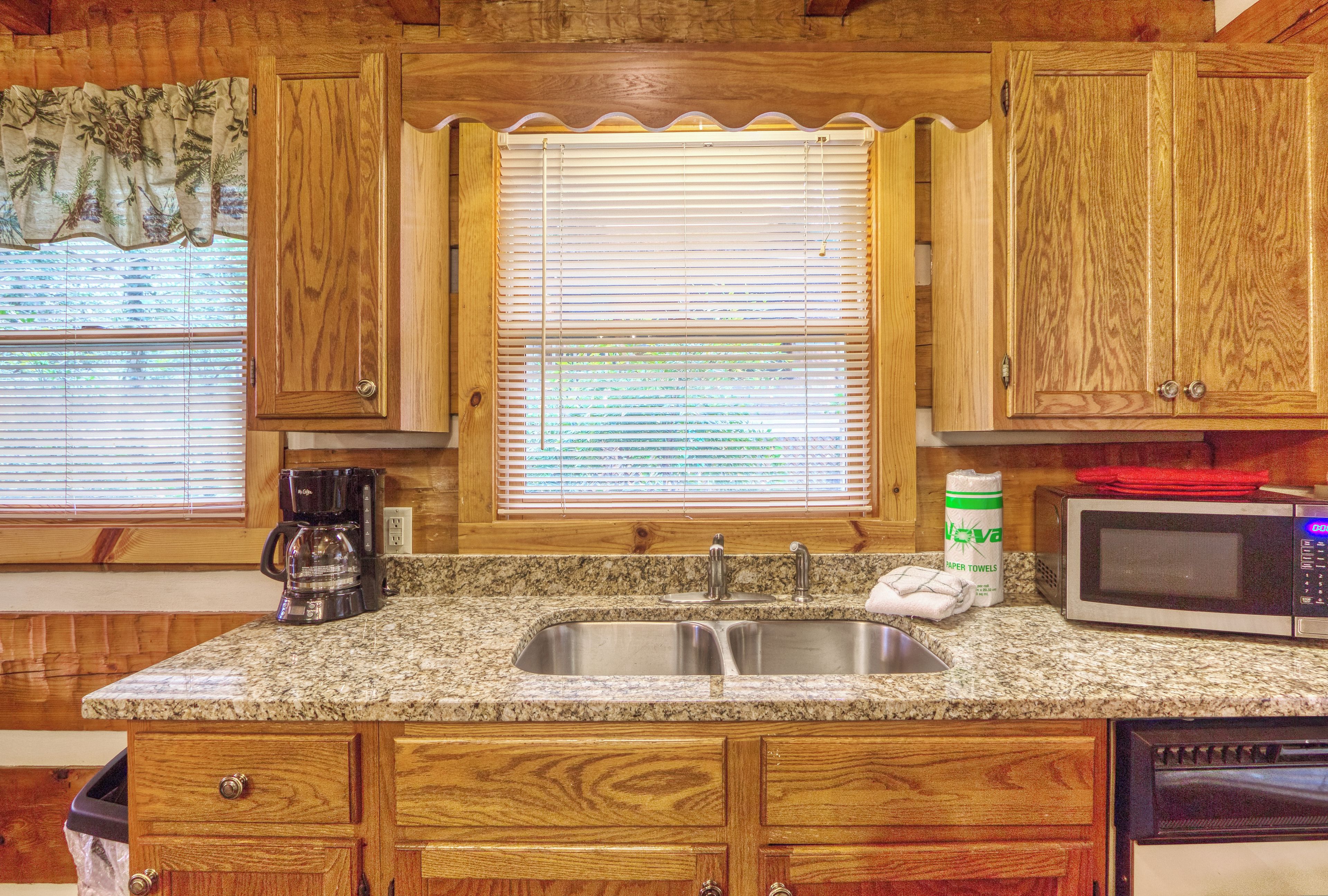 A kitchen with wooden cabinets, a granite countertop, a double sink, a coffee maker, a microwave, and a window with blinds.