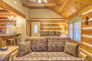 L’intérieur d’un chalet accueillant, avec un plafond en bois, un canapé agrémenté de coussins à motifs, un coin cuisine et une fenêtre équipée de stores.