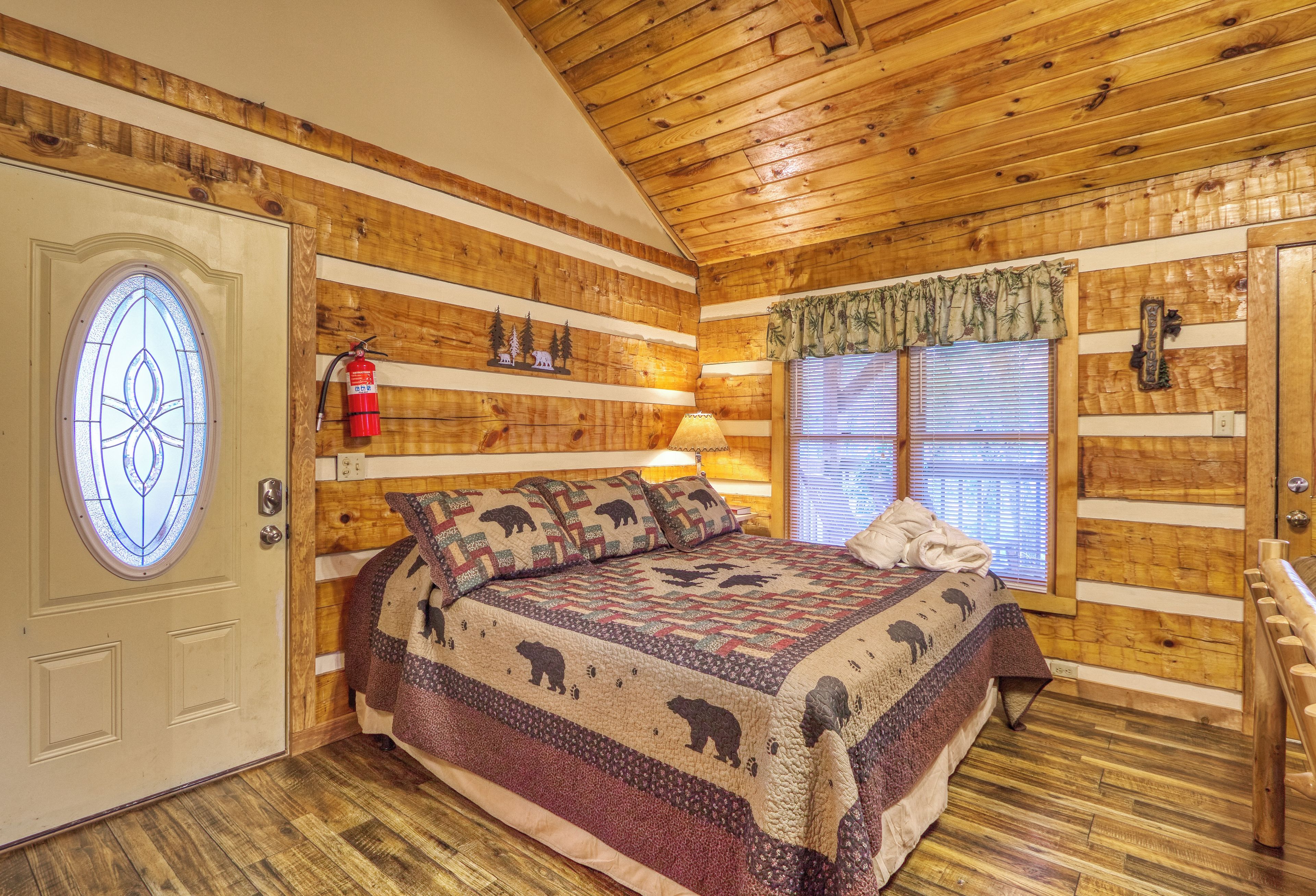 Une chambre dans une cabane en bois, avec un lit, une porte dotée d’une vitre, un extincteur et une fenêtre avec des stores.