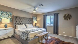 Um quarto com cabeceira estofada, uma cama grande, um banquinho, um ventilador de teto, uma janela com cortinas e um detalhe decorativo na parede.