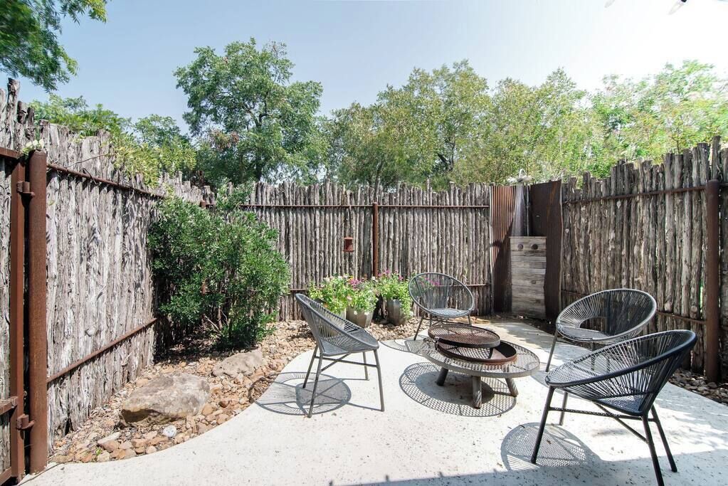 An outdoor patio area with a concrete floor, surrounded by a wooden fence. There are metal chairs and a round table with a central fire pit.