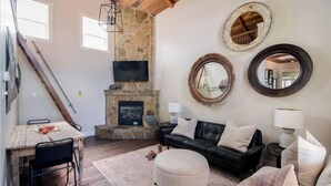 Un salon avec une cheminée en pierre, un canapé en cuir noir, une table à manger en bois et un escalier.