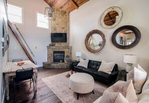 A living room with a stone fireplace, a black leather sofa, a wooden dining table, and a staircase. - Modern Guesthouse w/ Hot Tub + Fireplace (Fredericksburg)