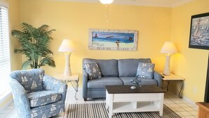 Un salon comprenant un canapé, un fauteuil et une table basse, un tableau représentant une scène de plage, ainsi que deux lampes posées sur des tables d’appoint.