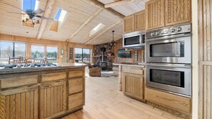 A spacious kitchen with wooden cabinets, a large island, and a stone fireplace. - Private Hot Tub! Game Room! Close to Slopes! Bright & open Amazing Views! (Big Bear City)