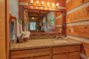 A bathroom with wooden paneling, a granite countertop, a sink, a mirror with lights, and a basket with towels.
