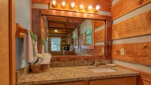 A bathroom with wooden paneling, a granite countertop, a sink, a mirror with lights, and a basket with towels. - Gatlinburg | Hot Tub | Pet Friendly | WiFi | Fireplace (GATLINBURG)