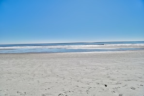 Una extensa playa de arena con huellas y un mar tranquilo bajo un cielo azul despejado.