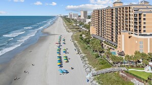 Un hotel frente a la playa con una extensa playa de arena, múltiples sombrillas y una zona de piscina.