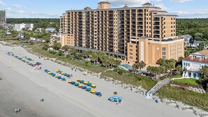 Un hotel grande con varios pisos, una playa con sombrillas y un sendero que lleva a la playa.