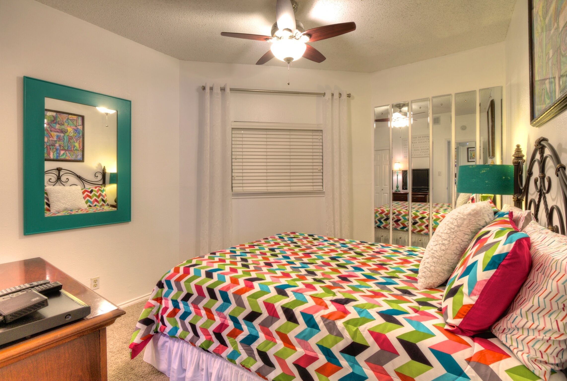A bedroom with a bed, a ceiling fan, a window with blinds, a bedside table, and a mirror.